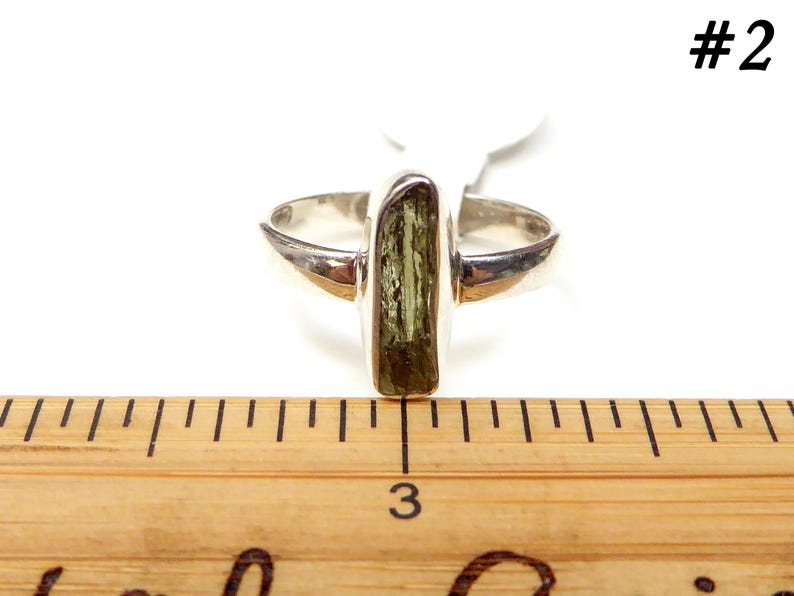 May include: A silver ring with a green and brown rectangular stone set in the center. The ring is shown on a wooden ruler with the number 3 visible.