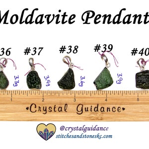 May include: Five green moldavite pendants on silver chains, each with a different shape and size. The pendants are displayed on a wooden ruler with measurements in inches. The text "#36", "#37", "#38", "#39", and "#40" are printed above each pendant. The text "Crystal Guidance" is printed on the ruler. The text "@crystalguidance stitchesandstoneskc.com" is printed below the ruler.