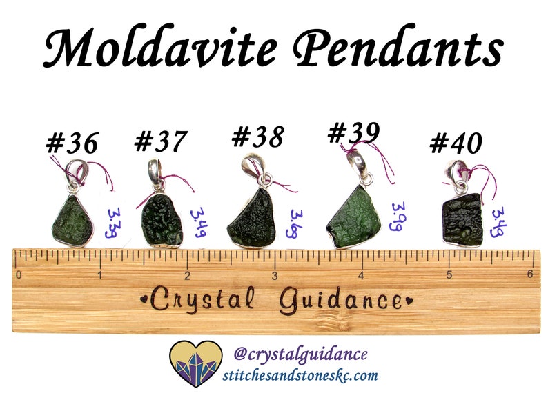 May include: Five green moldavite pendants on silver chains, each with a different shape and size. The pendants are displayed on a wooden ruler with measurements in inches. The text "#36", "#37", "#38", "#39", and "#40" are printed above each pendant. The text "Crystal Guidance" is printed on the ruler. The text "@crystalguidance stitchesandstoneskc.com" is printed below the ruler.