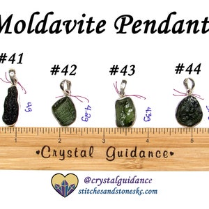 May include: Four Moldavite pendants with silver bail settings. The pendants are displayed on a wooden ruler with measurements in inches. The pendants are numbered #41, #42, #43, and #44. The text "Crystal Guidance" is printed on the ruler. The text "@crystalguidance stitchesandstoneskc.com" is printed below the ruler.