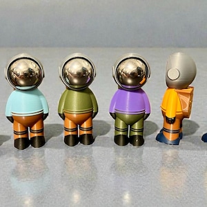 May include: A collection of six small figurines featuring astronaut-style helmets with reflective silver visors. The figures include a brown dog-like character, three standing figures in light blue, olive green, and purple, and two in yellow and orange.