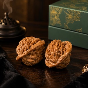 May include: Two brown, textured walnut shells with intricate, deeply grooved surfaces. The shells are positioned side-by-side, displaying a natural, organic appearance with a polished finish, highlighting the detailed ridges and patterns of the walnut exterior.