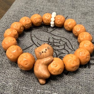 May include: A stretch bracelet featuring round, tan-colored beads embossed with small paw prints. The bracelet includes a central wooden charm shaped like a cat with a carved face and a small cluster of white spherical beads near the top section.