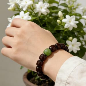 May include: A dark brown beaded bracelet with a single green bead. The bracelet is worn on a wrist near a potted plant with white flowers. The bracelet is made of dark brown beads and a single green bead.