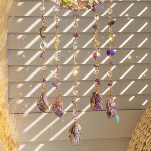May include: A decorative hanging crystal mobile featuring amethyst stones, clear and purple beads, and gold-colored crescent moon and star charms. The mobile is suspended by thin copper wire and hangs in front of a window with sunlight.