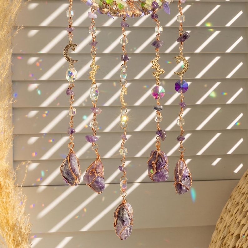 May include: A decorative hanging crystal mobile featuring amethyst stones, clear and purple beads, and gold-colored crescent moon and star charms. The mobile is suspended by thin copper wire and hangs in front of a window with sunlight.