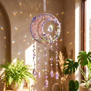 May include: A crescent moon-shaped dream catcher with pink and purple floral accents and hanging crystal beads. The dream catcher is suspended by a thin chain and features a large, teardrop-shaped crystal in the center. Decorative celestial-themed home decor.
