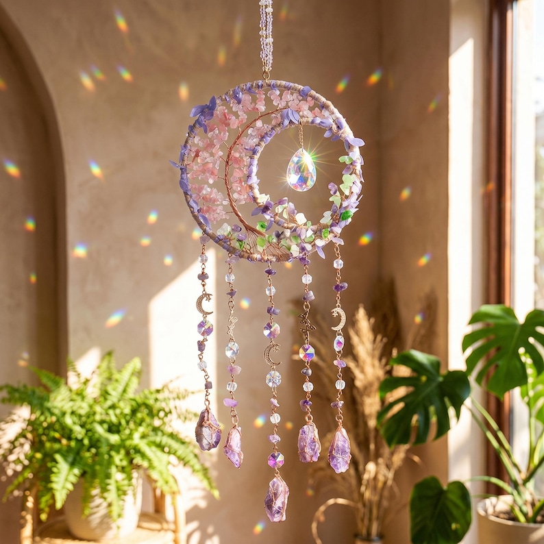 May include: A crescent moon-shaped dream catcher with pink and purple floral accents and hanging crystal beads. The dream catcher is suspended by a thin chain and features a large, teardrop-shaped crystal in the center. Decorative celestial-themed home decor.