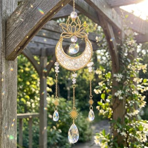 May include: A gold-toned metal suncatcher featuring a lotus flower top, a crescent moon filled with clear crystals, and three hanging chains. The chains are adorned with small clear crystal beads, gold sun charms, and faceted clear crystal pendants.