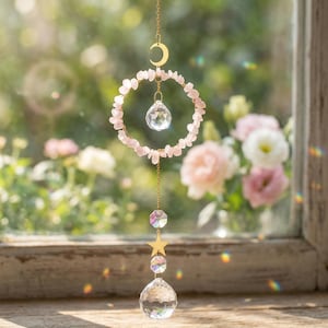 May include: A hanging suncatcher with a gold-colored crescent moon and chain. A circle of pink stones surrounds a faceted crystal. Below, a gold star and two more crystals hang from the chain. The background is a blurred view of flowers.