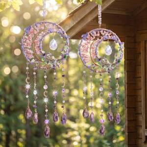 May include: Two celestial-themed hanging decorations. Each features a crescent moon shape adorned with pink, purple, and green crystals. Clear teardrop-shaped crystals hang from the center, with strands of beads and amethyst crystals dangling below. The decorations are suspended from a wooden structure.