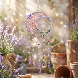 May include: A celestial-themed dream catcher featuring a crescent moon design. The moon is adorned with pink and purple flowers, green accents, and a dangling crystal. Strands of beads and amethyst crystals hang below, with gold-colored moon charms.