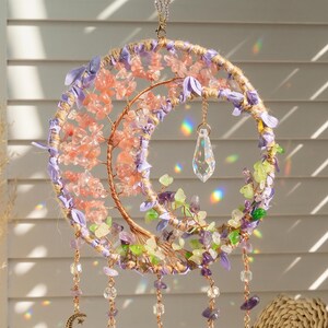 May include: A handcrafted dream catcher with a crescent moon design. It features a copper wire tree adorned with pink and green gemstones, surrounded by a woven frame with purple accents. A clear crystal pendant hangs from the center, with dangling gemstone strands.
