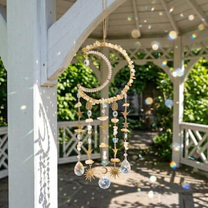 May include: A celestial-themed suncatcher with a crescent moon design. The gold-toned metal frame is adorned with small, light-colored stones and dangling crystal accents. Additional charms include stars, suns, and teardrop-shaped crystals. The suncatcher hangs from a delicate chain.