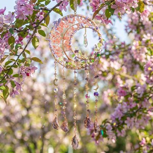 May include: A celestial-themed suncatcher with a crescent moon design. The moon is adorned with pink and purple crystals and gemstones. Dangling from the moon are strands of beads, crystals, and amethyst stones. The suncatcher hangs from a gold chain.