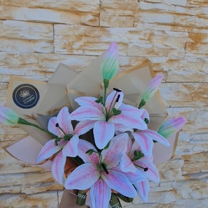 Puede incluir: Un ramo de lirios rosas y blancos con detalles y capullos verdes, envuelto en papel marrón. El ramo está atado con una cinta verde que dice "Just for you". El fondo es una pared texturizada de color marrón claro.