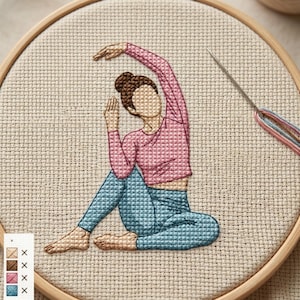 Puede incluir: Bordado de punto de cruz de una persona en una postura de yoga, con una parte superior rosa y pantalones azules. El diseño está en un bastidor de bordado de madera con aguja e hilo visibles. La paleta de colores incluye rosa, azul, marrón y beige.