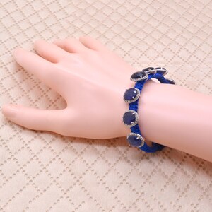 May include: A bracelet featuring a vibrant blue braided cord adorned with multiple round, dark blue cabochon stones. Each stone is set in a silver-toned metal frame with small, clear, sparkling accents along the outer edge of the setting.