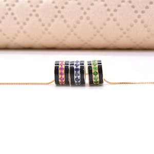 Op de afbeelding: Een gouden ketting met drie cilindervormige kralen. Elke kraal heeft een zwarte emaille afwerking met een centrale band van kleine, ronde, gezette edelstenen in roze, blauw en groen, horizontaal gerangschikt op de dunne gouden ketting.
