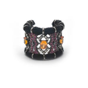 Può includere: Un anello a fascia larga con una struttura in metallo scuro avvolta da un cordino in fibra nera. Il design presenta gemme ovali arancioni incastonate in un motivo a reticolo, arricchito da piccole pietre rosa e bianche nel metallo.