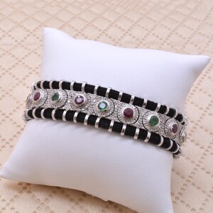 May include: A bracelet featuring a central silver-toned band with alternating round red and green gemstones set in textured metal. The central band is flanked by two black cord sections held in place by evenly spaced silver-toned metal loops.