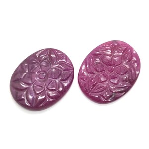 May include: Two oval-shaped, deep magenta-colored gemstones featuring a carved floral pattern on their surfaces. Each stone displays a symmetrical, engraved design with petal-like motifs radiating from a central circular point, polished to a smooth finish.