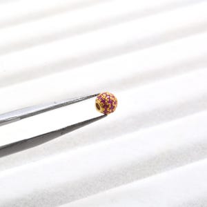 May include: A small, spherical gold bead featuring a textured surface embedded with numerous tiny, vibrant pink stones. The bead is held by a pair of metal tweezers, showcasing its intricate design and circular shape for jewelry making.