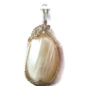 May include: A pear-shaped pendant with a large, iridescent, cream-colored stone. The stone is encased in a silver-toned metal frame with a decorative, filigree design at the top. The pendant hangs from a silver-toned bail.