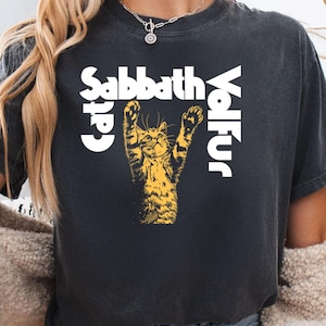 May include: Black t-shirt featuring a yellow illustration of a cat with its paws raised. The text "Sabbath Cat Volfur" is printed in white, with "Cat" and "Volfur" vertically aligned on either side of the cat image.