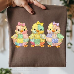 May include: A brown sweatshirt featuring three colorful patchwork chick designs. Each chick has a bow and is made of various fabric squares in shades of yellow, pink, green, and purple. The chicks have orange beaks and feet.
