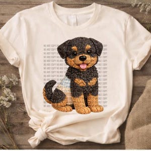 Peut inclure: T-shirt crème avec une illustration détaillée d'un chiot Rottweiler. Le chiot a des poils noirs et bruns, une langue rose et est assis. Le t-shirt porte le texte "DO NOT COPY" répété en arrière-plan.