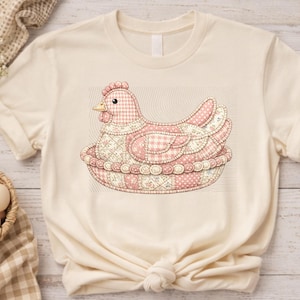 Pode incluir: Camiseta creme com uma ilustração detalhada de uma galinha de patchwork rosa e branco em um ninho. A galinha tem uma crista e um bico vermelhos, com vários tecidos estampados usados no design. O ninho também é de patchwork.