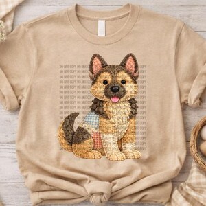 Puede incluir: Camiseta color beige con una ilustración detallada de un perro pastor alemán. El perro está representado con pelaje marrón, negro y beige, sentado con la lengua fuera. La camiseta tiene cuello redondo y mangas cortas.