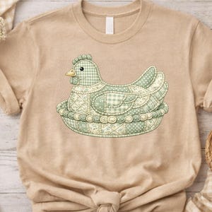 May include: A tan-colored t-shirt featuring a detailed illustration of a hen sitting in a nest. The hen and nest are in shades of green and white, with a patchwork quilt design. The hen has a yellow beak and a decorative comb.
