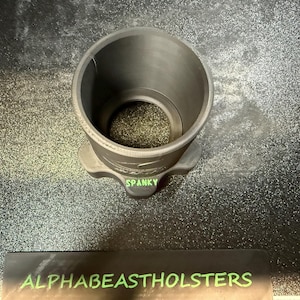 Puede incluir: Un objeto cil&iacute;ndrico gris oscuro con una base ensanchada, etiquetado "SPANKY" en texto verde. El objeto se encuentra sobre una superficie texturizada. La parte inferior de la imagen muestra un rect&aacute;ngulo negro con el texto "ALPHABEASTHOLSTERS" en verde.