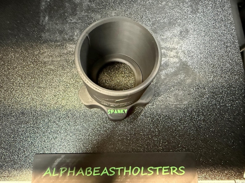 Puede incluir: Un objeto cil&iacute;ndrico gris oscuro con una base ensanchada, etiquetado "SPANKY" en texto verde. El objeto se encuentra sobre una superficie texturizada. La parte inferior de la imagen muestra un rect&aacute;ngulo negro con el texto "ALPHABEASTHOLSTERS" en verde.