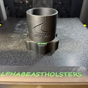 Puede incluir: Un portavasos negro impreso en 3D con el logotipo de Ski-Doo y la palabra "SPANKY" en verde. La base tiene una forma &uacute;nica. El texto "ALPHABEASTHOLSTERS" est&aacute; impreso en verde sobre una base rectangular negra.