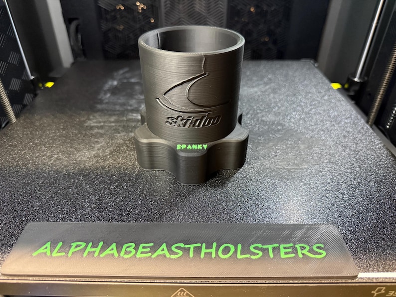 Puede incluir: Un portavasos negro impreso en 3D con el logotipo de Ski-Doo y la palabra "SPANKY" en verde. La base tiene una forma &uacute;nica. El texto "ALPHABEASTHOLSTERS" est&aacute; impreso en verde sobre una base rectangular negra.