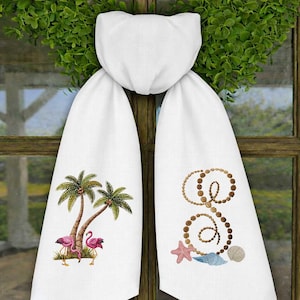 May include: White decorative towel set with embroidered tropical designs. One towel features palm trees and pink flamingos, the other has a beaded letter "S" with seashells and a starfish. The towels are hanging on a wooden window.