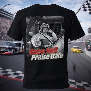 Puede incluir: Camiseta negra con una imagen en blanco y negro de un piloto de carreras haciendo una peineta. El texto "Raise Hell Praise Dale" está impreso en rojo y blanco. Coches de carreras y una bandera a cuadros en el fondo.