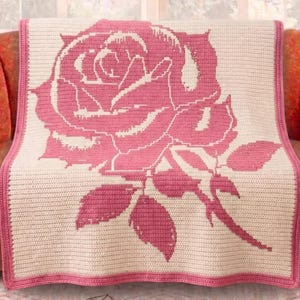 Puede incluir: Una manta de ganchillo con un gran diseño de rosa rosa sobre un fondo crema. La manta tiene un borde rosa y está colocada sobre un sofá naranja. La rosa tiene pétalos y hojas detallados, creando un textil para el hogar acogedor y decorativo.