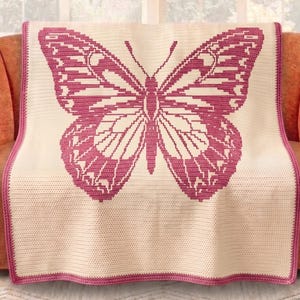May include: A cream-coloured throw blanket featuring a large magenta butterfly design. The butterfly is centrally placed with detailed wing patterns. The blanket has a magenta border and is draped over an orange sofa.
