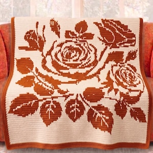 May include: A cream-colored crocheted blanket with a large rose design in shades of brown. The blanket is draped over an orange sofa, showcasing the intricate floral pattern and detailed edging. The blanket's dimensions are approximately 60 inches by 50 inches.