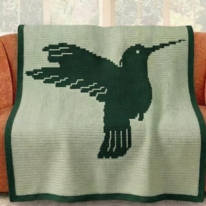 May include: A light green and dark green crocheted blanket featuring a large hummingbird design. The blanket has a dark green border and is draped over an orange sofa. The hummingbird is depicted in a silhouette style.