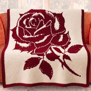 May include: A cream-colored crocheted blanket with a large, detailed burgundy rose design. The blanket has a burgundy border and is draped over an orange sofa. The rose features intricate petal and leaf details.