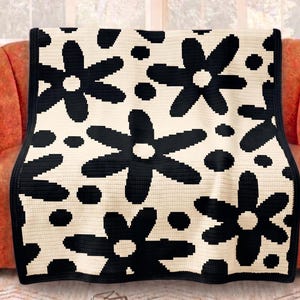May include: A cream-colored crocheted blanket with a repeating pattern of large black flowers and black dots. The blanket is draped over an orange sofa, creating a cozy and decorative accent for a living space.