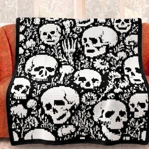 Pode incluir: Um cobertor preto e branco com um padrão repetido de caveiras. As caveiras são brancas sobre um fundo preto, criando um design de alto contraste. O cobertor está sobre um sofá laranja.