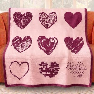 May include: A pink crocheted blanket with nine heart designs in varying styles and shades of burgundy. The blanket is draped over an orange sofa, with a window in the background. The blanket has a burgundy border.