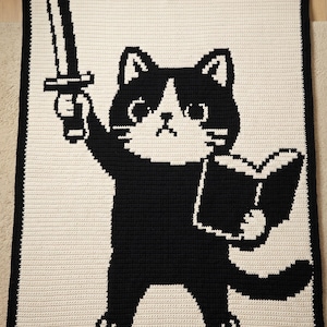 May include: A rectangular crochet blanket featuring a black and white graphic design of a cat standing on its hind legs. The cat holds a sword raised in one paw and an open book in the other. The blanket has a solid black border on a cream background.