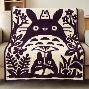 May include: A rectangular crochet blanket featuring a dark purple and cream colour scheme. The design displays a large central creature with long ears, a smaller creature on its head, and a third creature holding a leaf umbrella below.